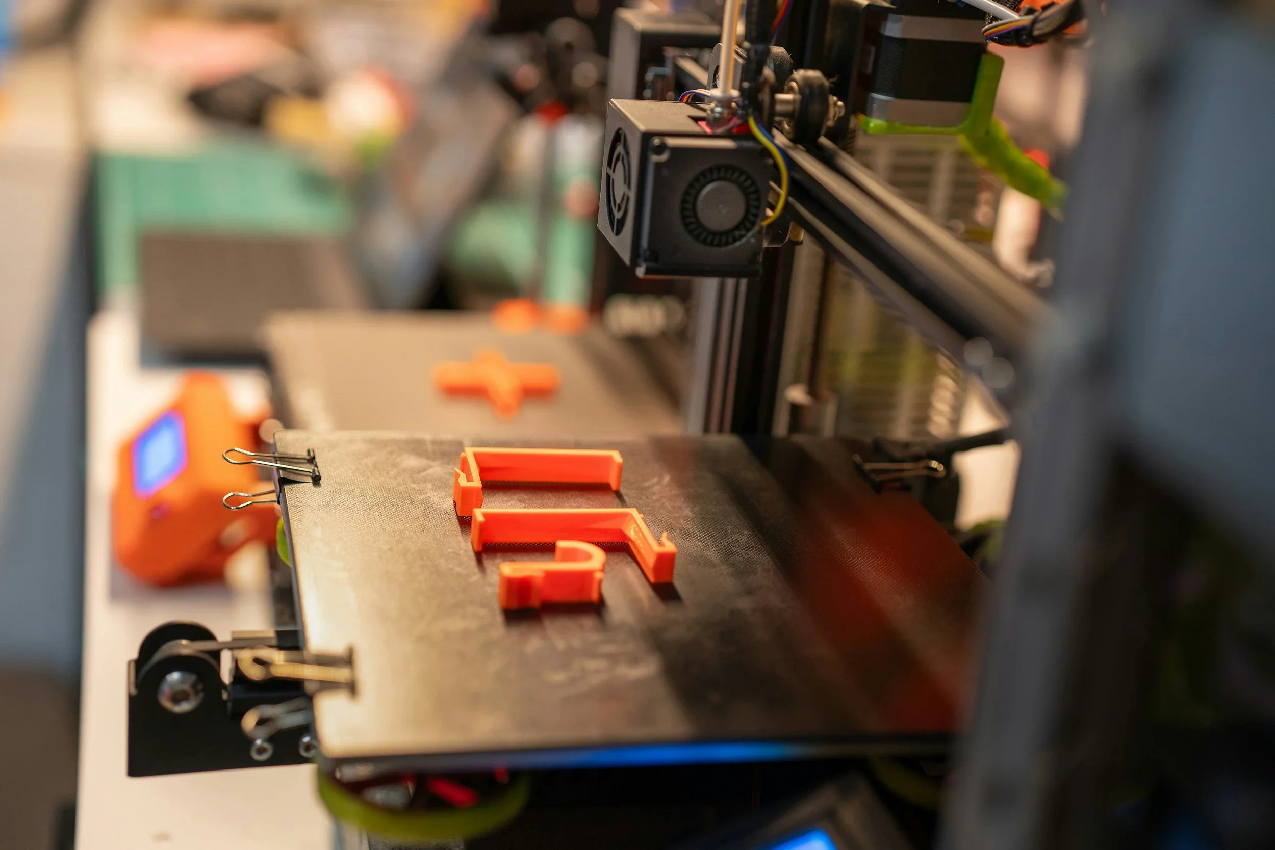 A 3D printer creating red plastic parts on a build platform in a workshop setting with various components and tools visible in the background.