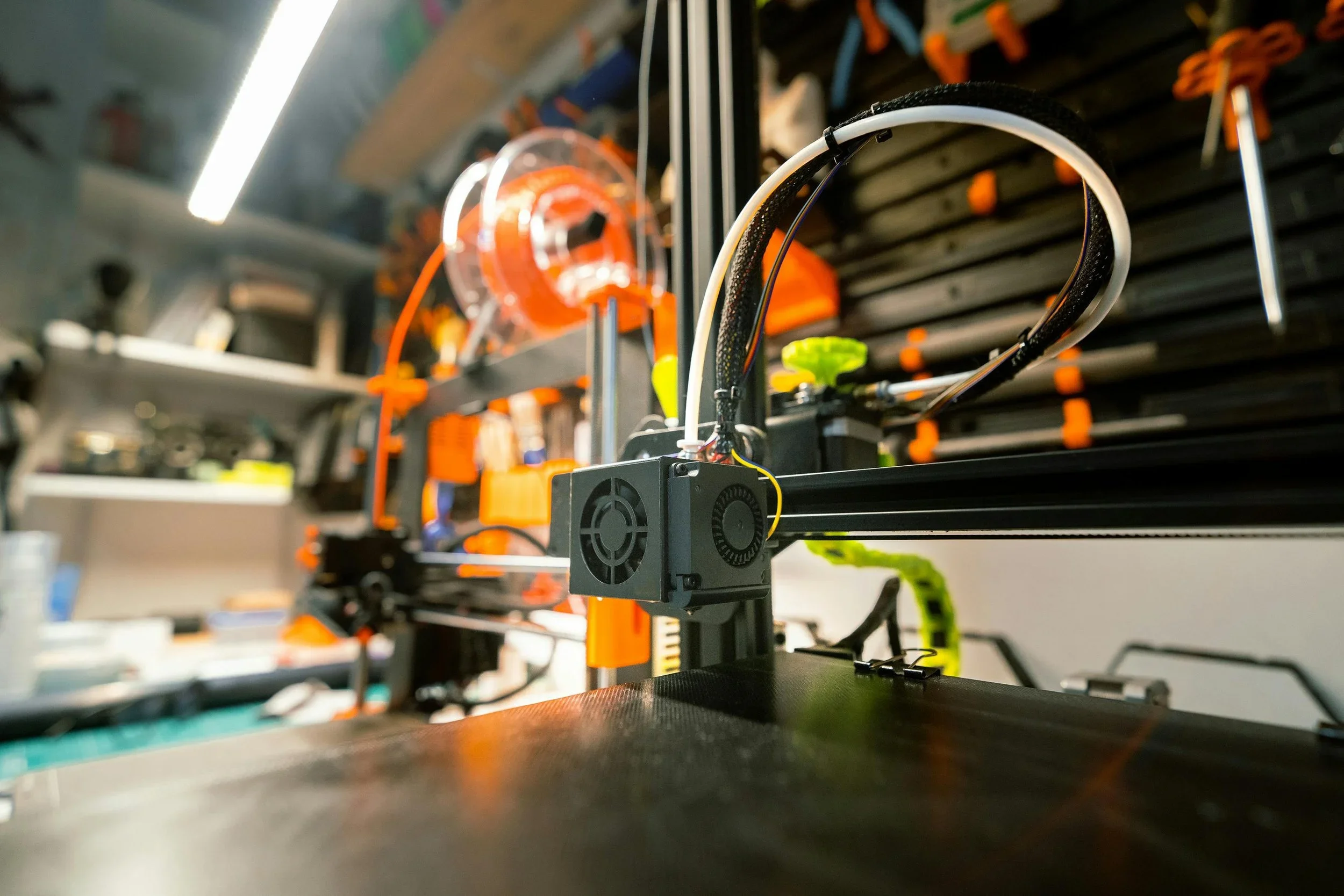 3D printers with orange and black frames in a workshop showroom with fluorescent lighting and workstations in the background.