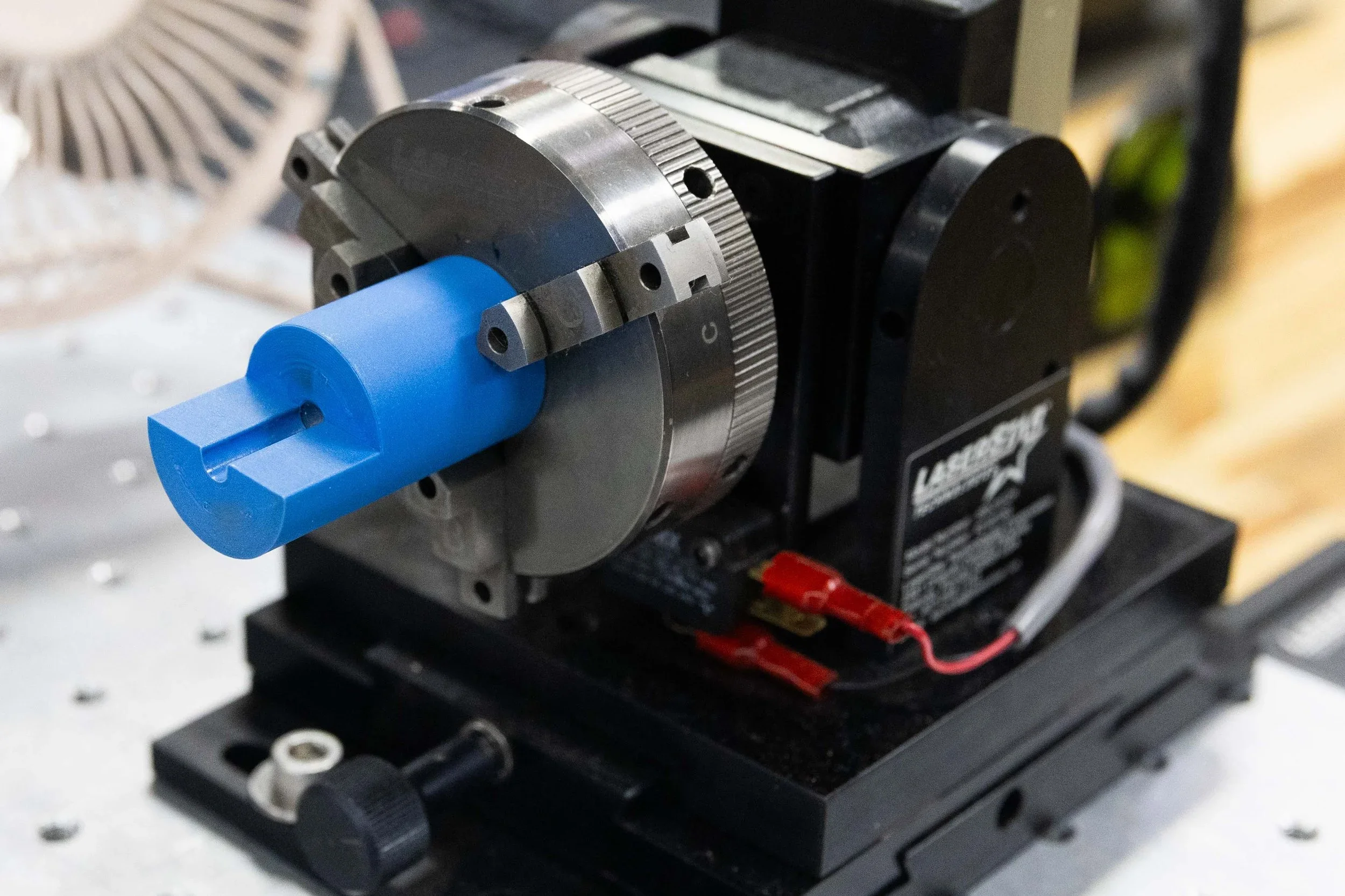 A blue motorized roller shade mechanism with metal gearing and control components mounted on a black base, displayed in a showroom or workshop setting.