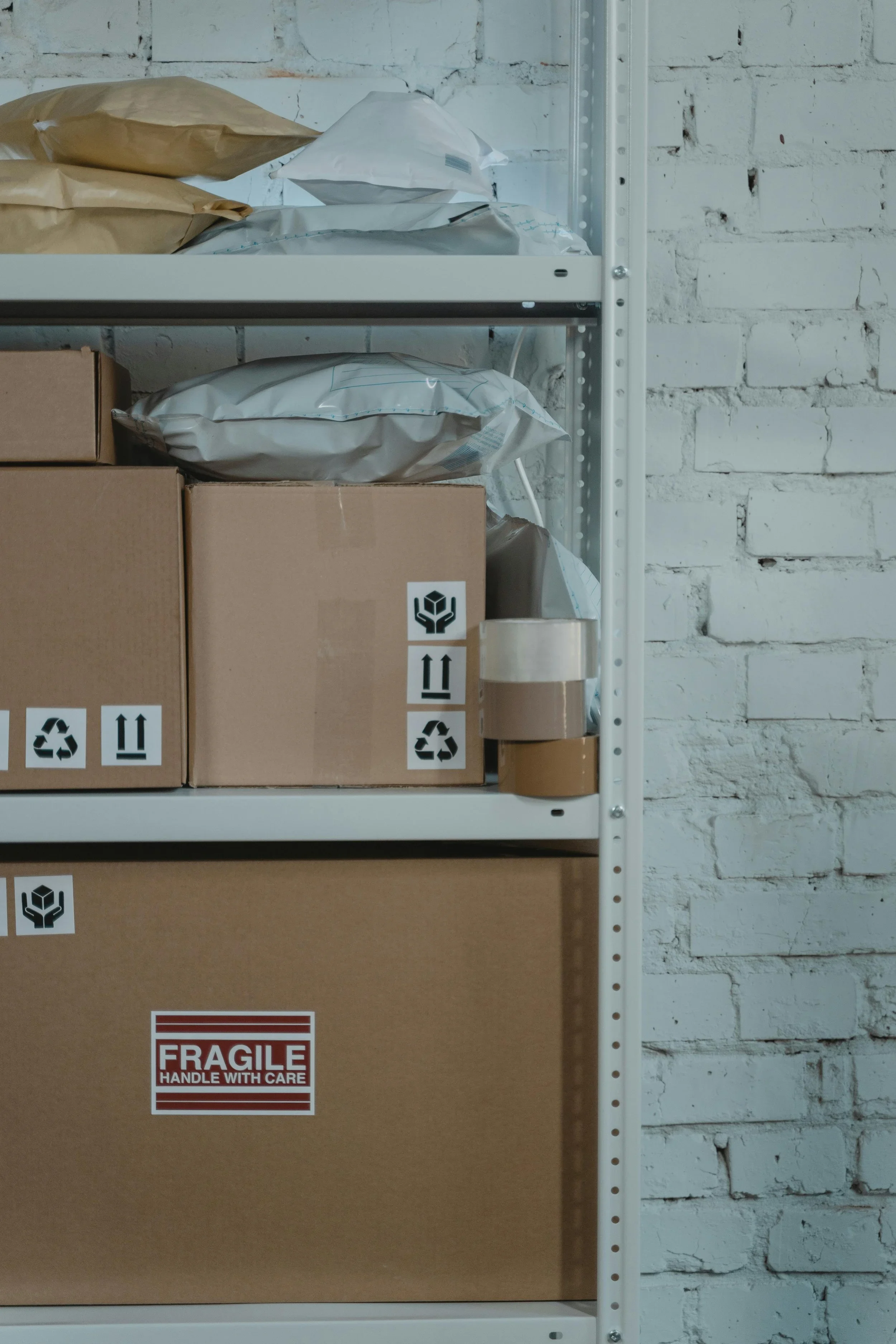 Cardboard shipping boxes with fragile and recycling labels stored on metal industrial shelving against a white brick wall.