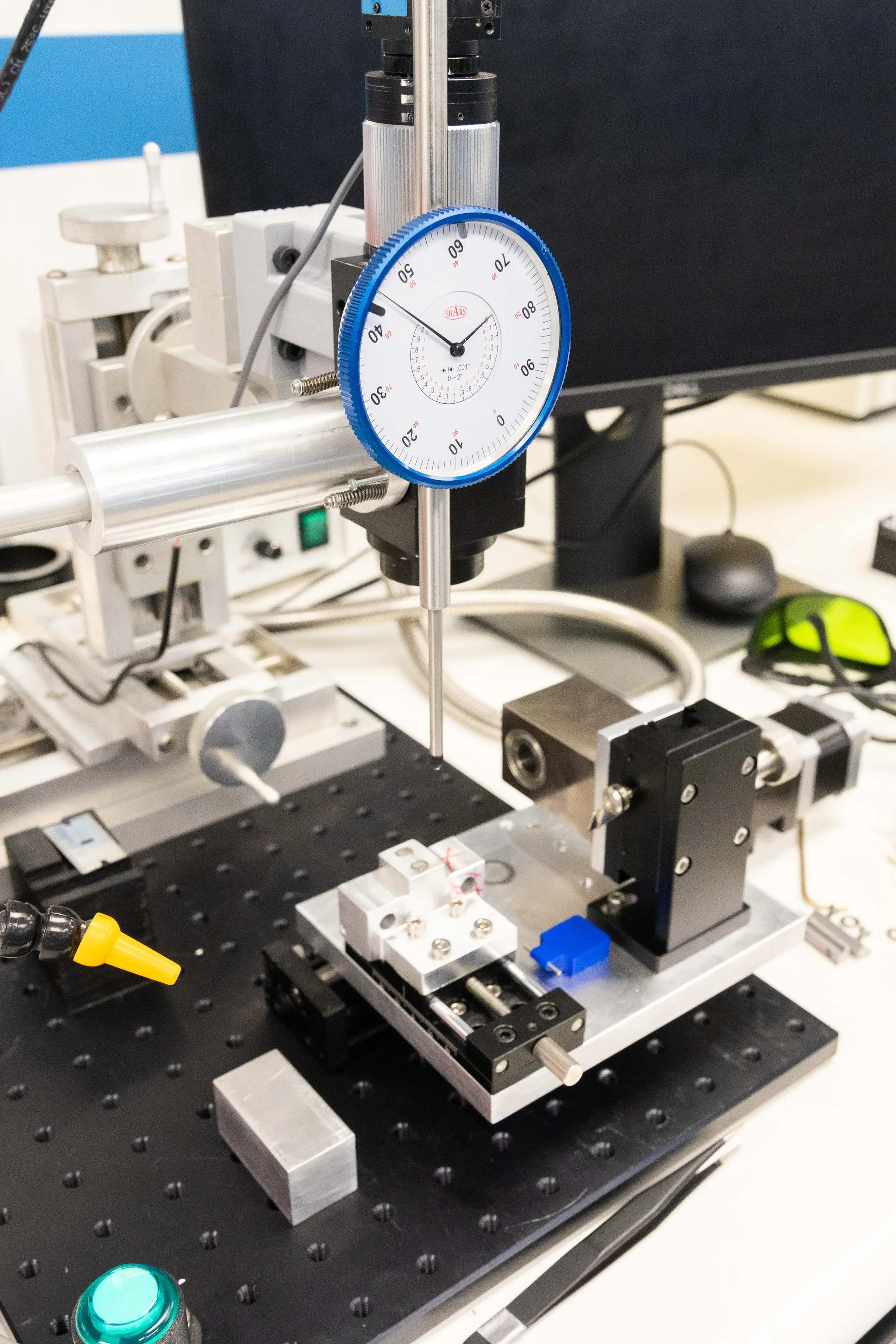 A dial gauge measurement instrument mounted on a precision testing apparatus with electronic components on an optical breadboard in a laboratory setting.
