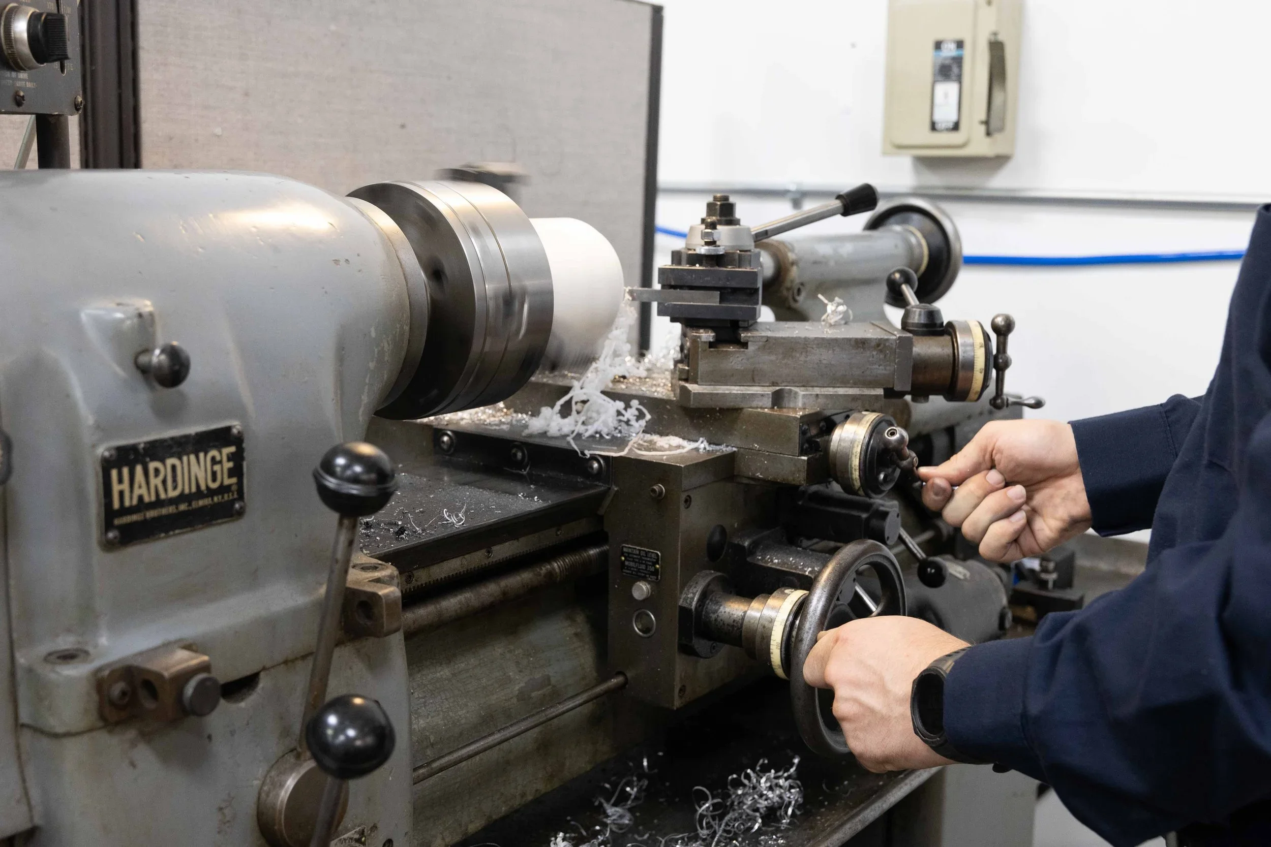 A Hardinge lathe with metal workpiece and cutting tools being operated by hands in an educational or workshop setting.
