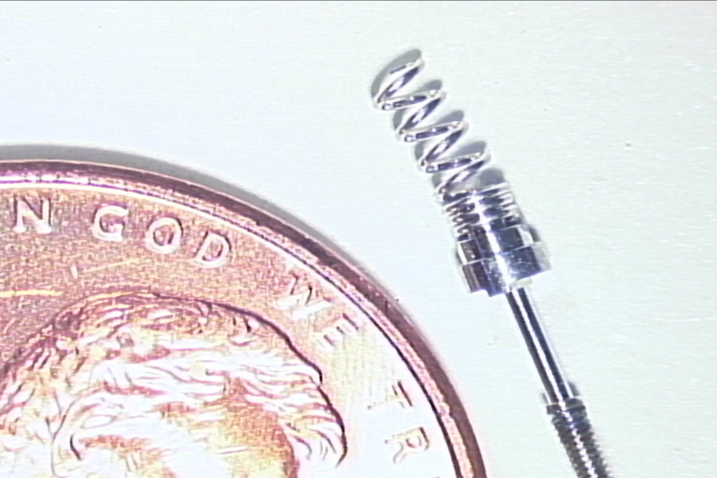 A mascara brush with spiral bristles displayed next to a decorative coin or medallion showing detailed metalwork and text.