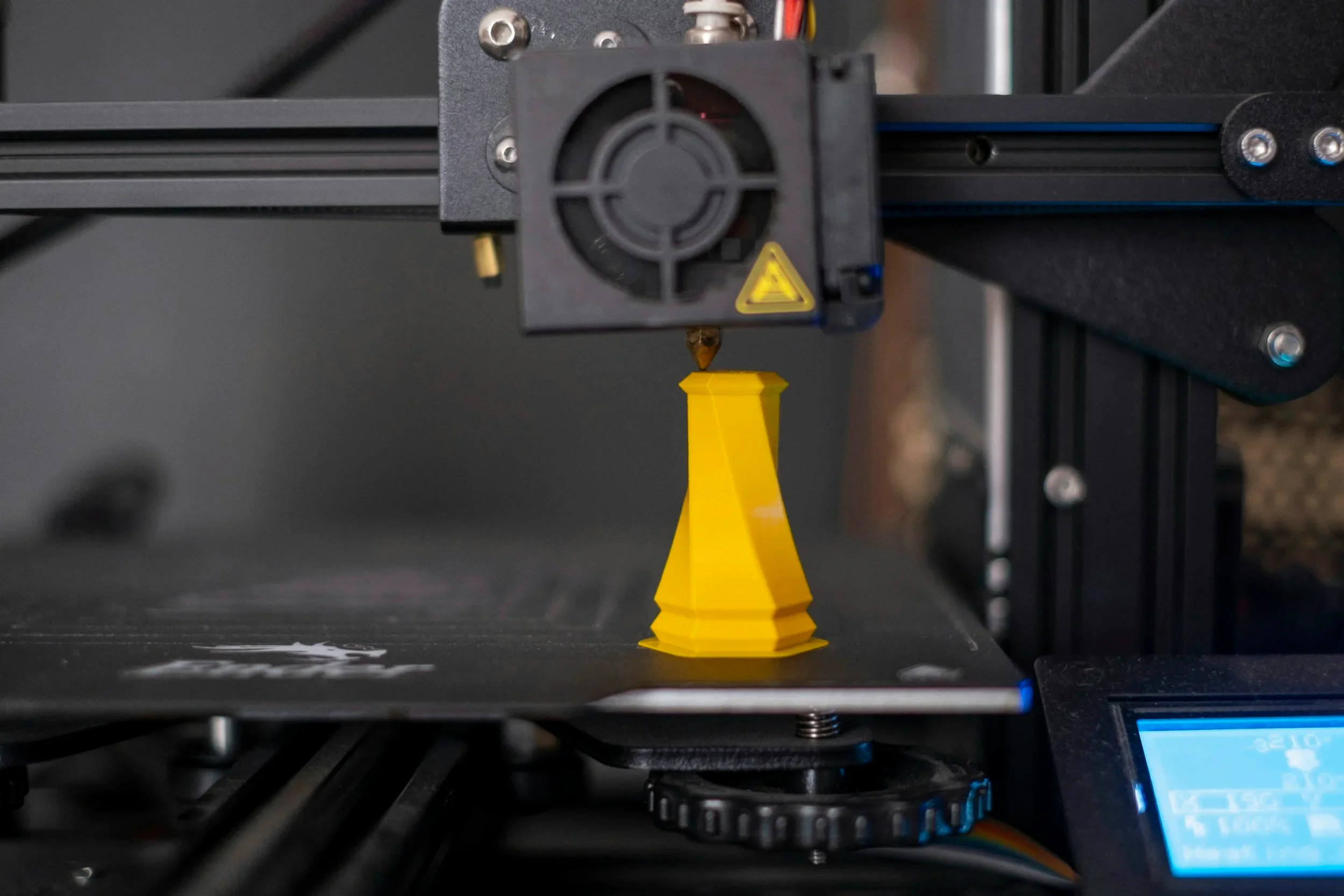 A yellow 3D-printed cone being produced by a desktop 3D printer on its build platform in a workshop setting.
