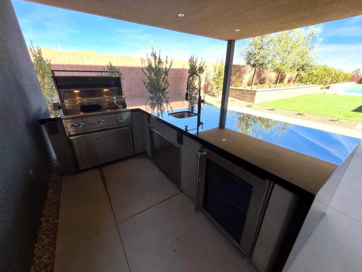 An outdoor kitchen with built-in grill and countertops in a covered patio area overlooking a manicured backyard.