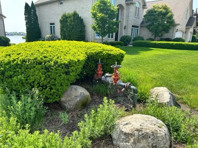 Outdoor water valve and piping installation in a residential landscape with decorative hedges and boulders.