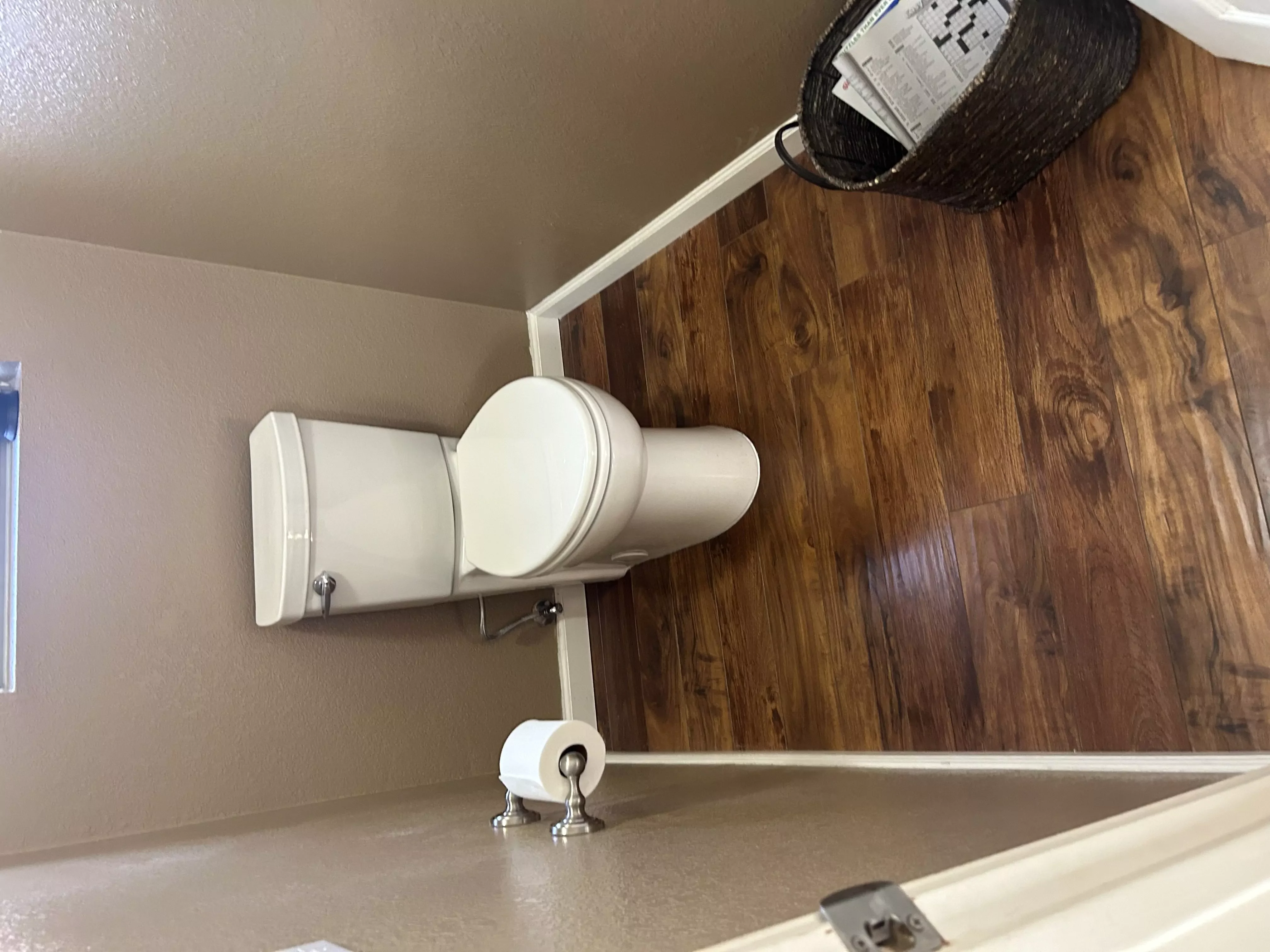 A toilet installation in a residential bathroom with wood flooring and beige walls.