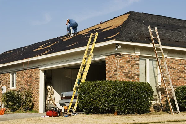 Roof replacement in progress on a brick residential home, with workers on ladders removing old shingles from the pitched roof.