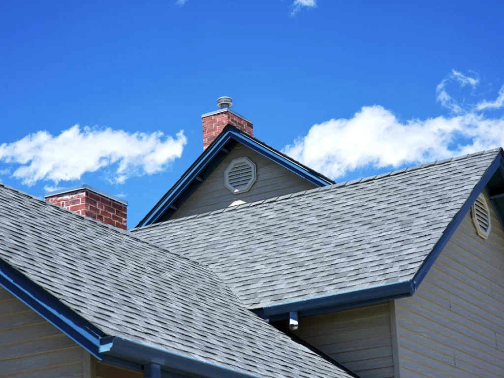 Asphalt shingle roof installation with multiple gables, brick chimneys, and blue trim trim details photographed against a blue sky.