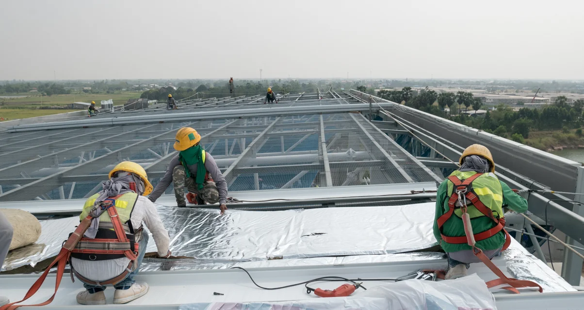Flat roof installation with workers applying protective membrane on a large commercial roof structure in a rural setting.