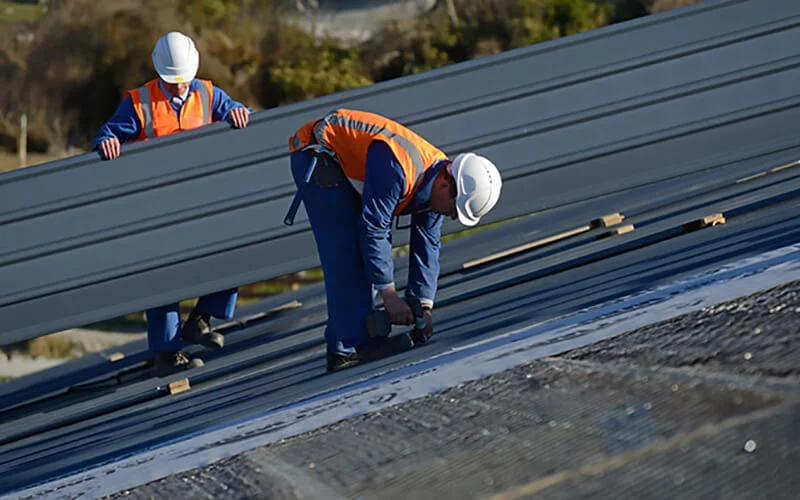 Metal roof installation on a residential roofline with two workers in safety gear securing corrugated metal panels.