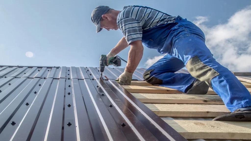 Metal roof installation with a worker using a power drill to secure corrugated metal panels onto wooden roof decking under clear sky conditions.