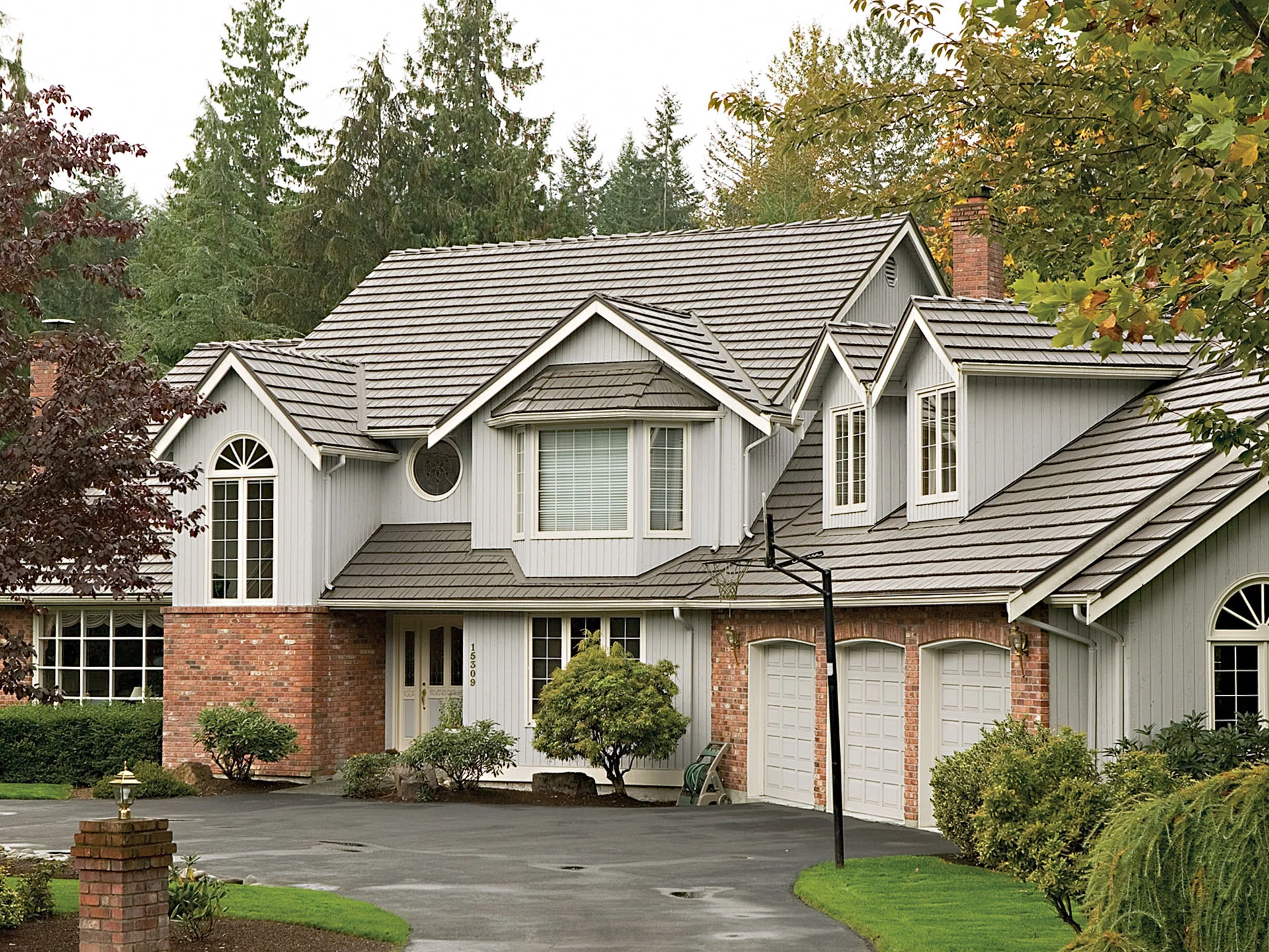 A residential metal/architectural shingle roof on a two-story brick and siding home with multiple roof peaks and dormers in a wooded suburban setting.