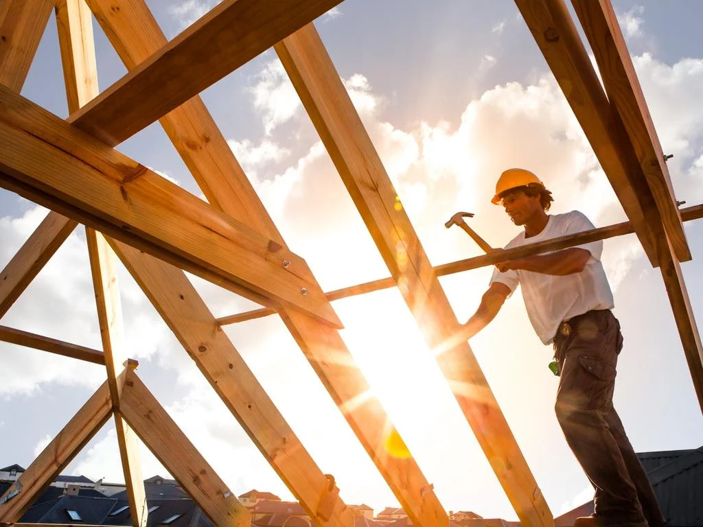Roof framing installation showing a worker with a hammer working on wooden roof trusses against a bright sky.
