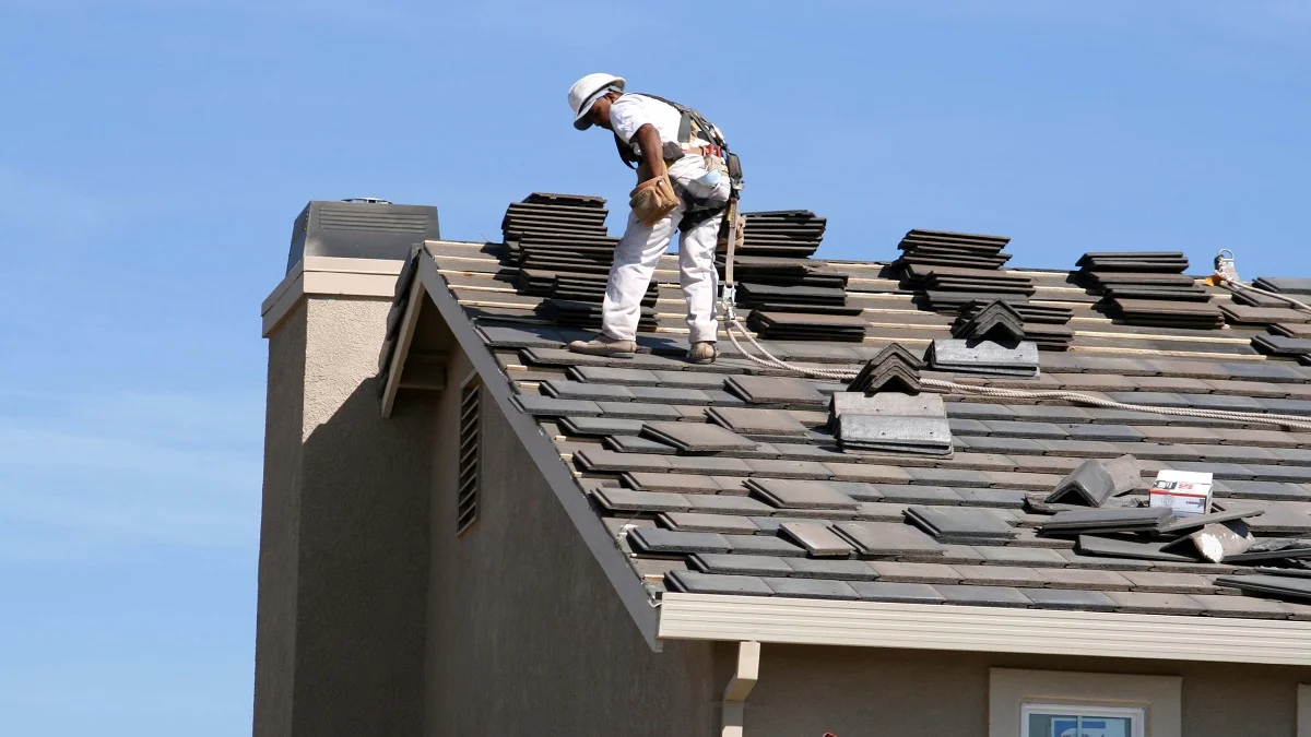 Roof installation with asphalt shingles being laid on a residential home during active construction, showing a worker on the roof against a clear blue sky.