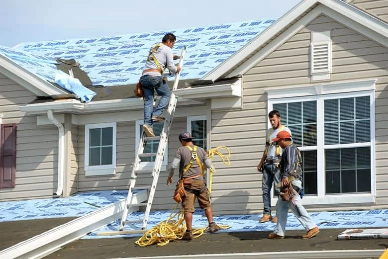 Roof installation with blue underlayment and shingles being applied by workers on a residential home during active construction.