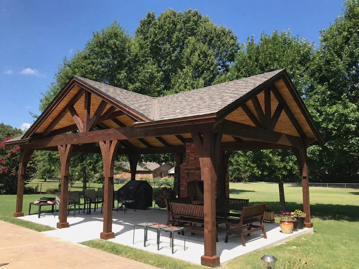A custom wooden pergola with gable roof structure and brick chimney over a concrete patio in a residential backyard setting.