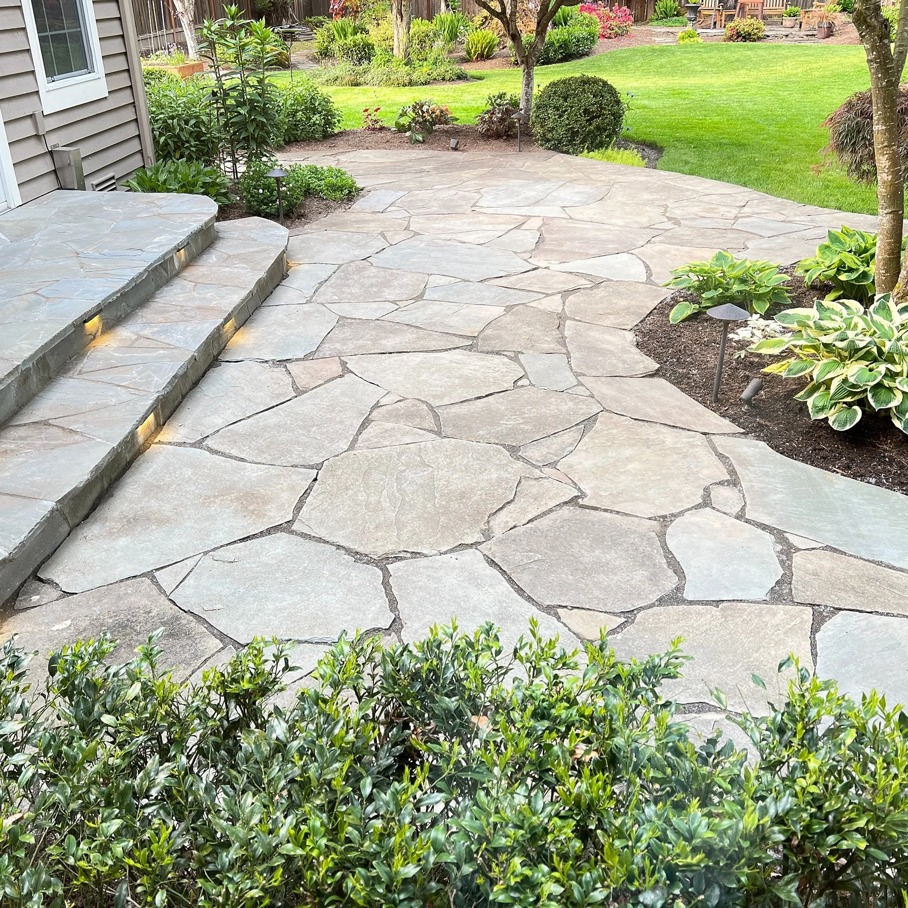 A flagstone patio installed in a residential backyard setting with landscaping and lawn.