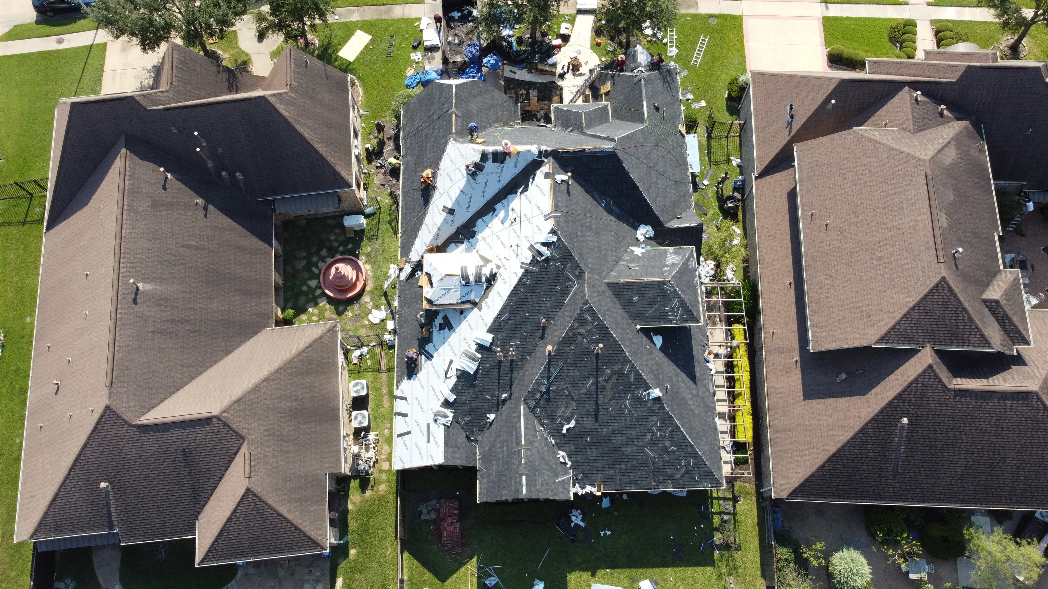 Roof replacement work in progress on a residential home, showing workers installing new shingles and protective coverings across the roof structure.