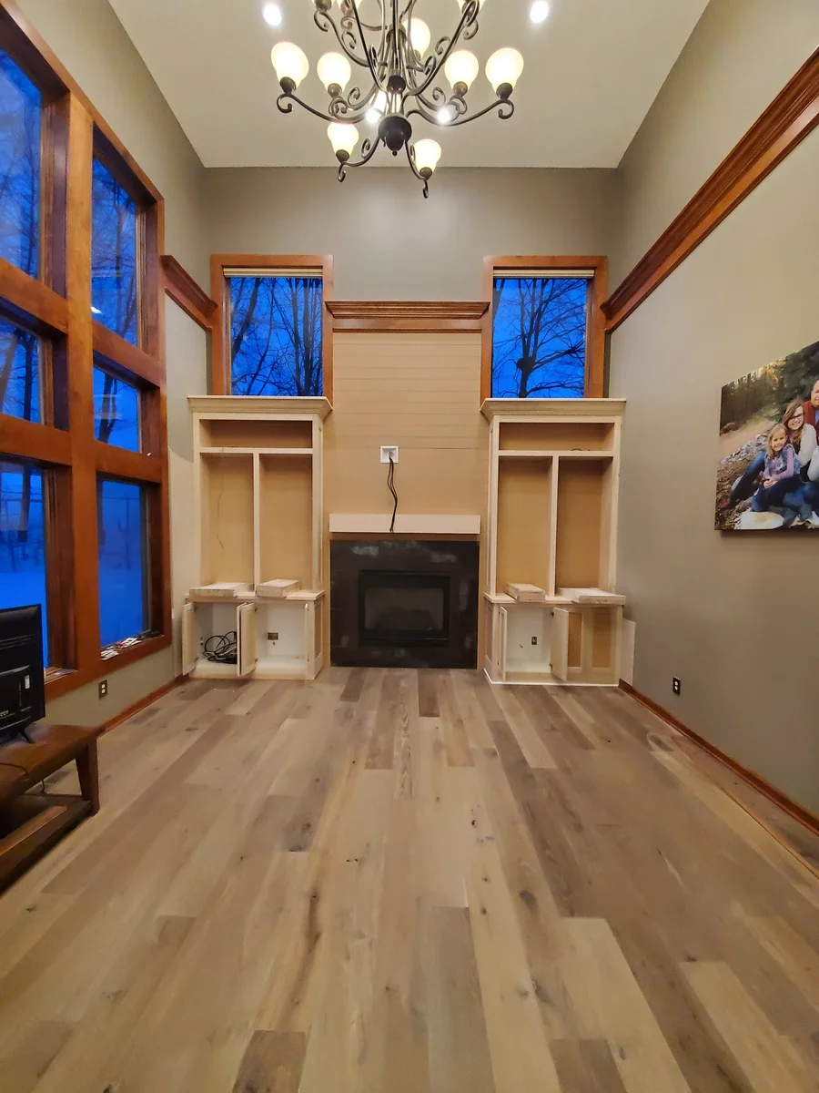 Built-in wooden shelving and cabinetry flanking a fireplace in a high-ceilinged living room with large windows and hardwood flooring.