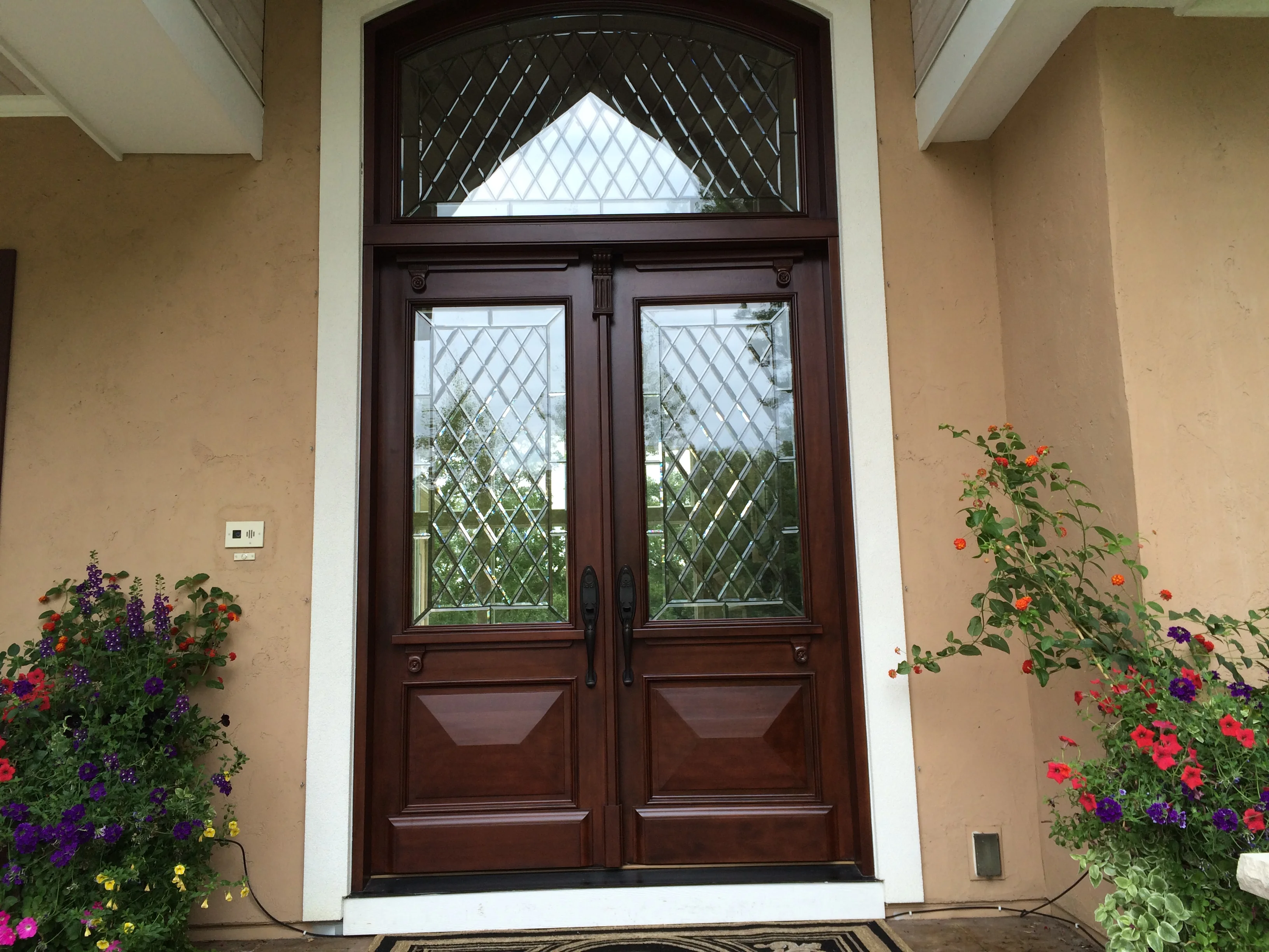 Double wooden entry doors with diamond-pattern glass panes and transom window in a cream-colored entryway flanked by flowering planters.