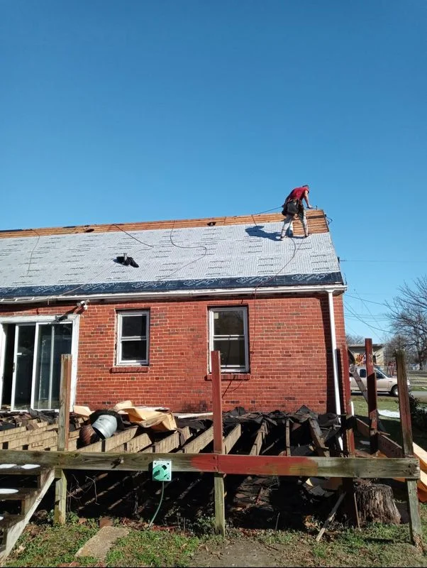 Roof replacement work in progress on a brick residential home, with a worker installing new roofing materials on the pitched roof.
