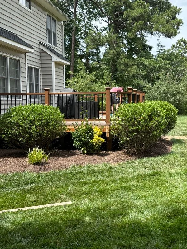 A wooden deck with black railings and integrated seating attached to a residential home, surrounded by landscaped bushes and lawn.