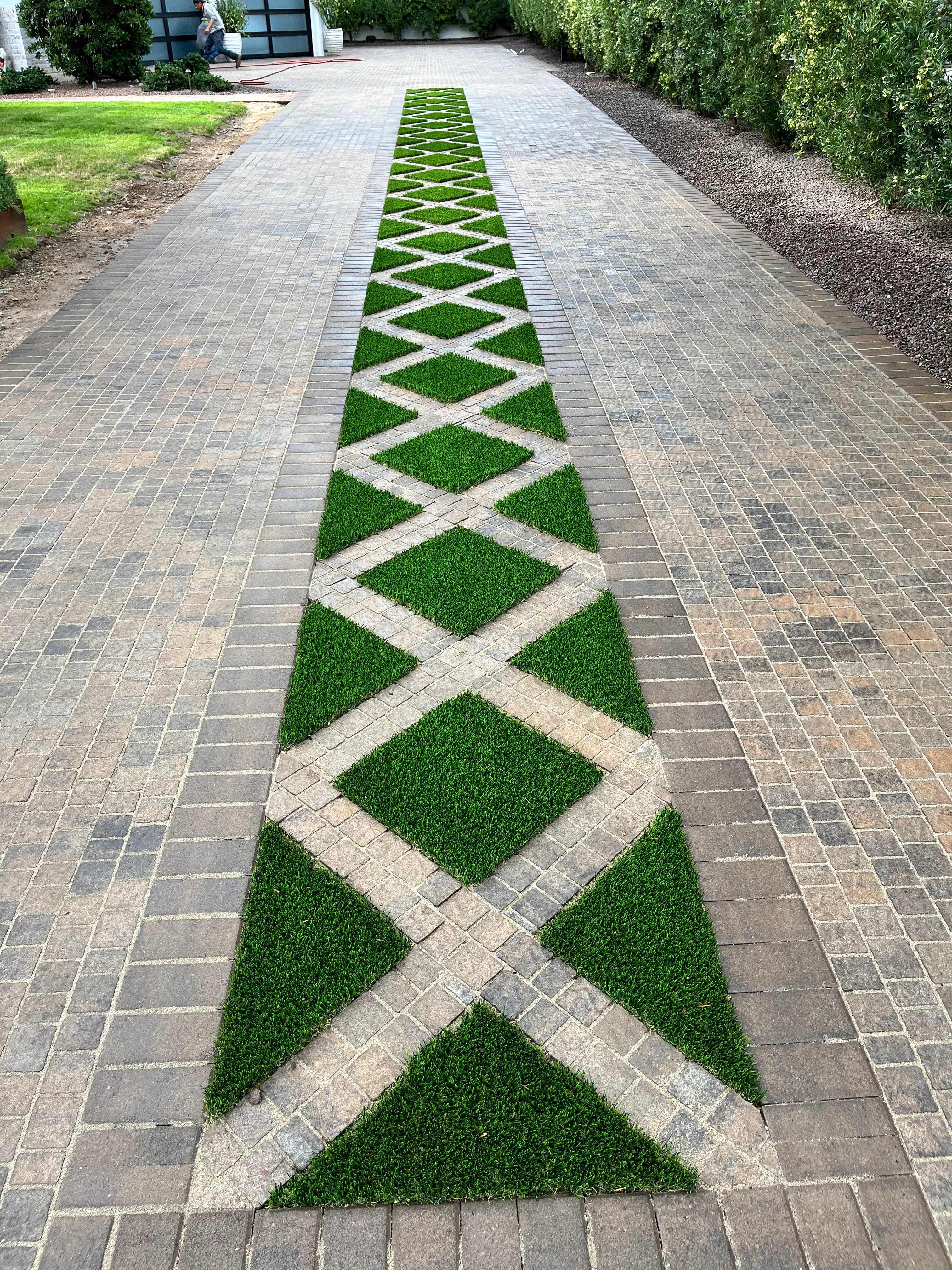 Decorative paver driveway with geometric grass inlay pattern featuring diamond and crosshatch designs in a residential landscape setting.