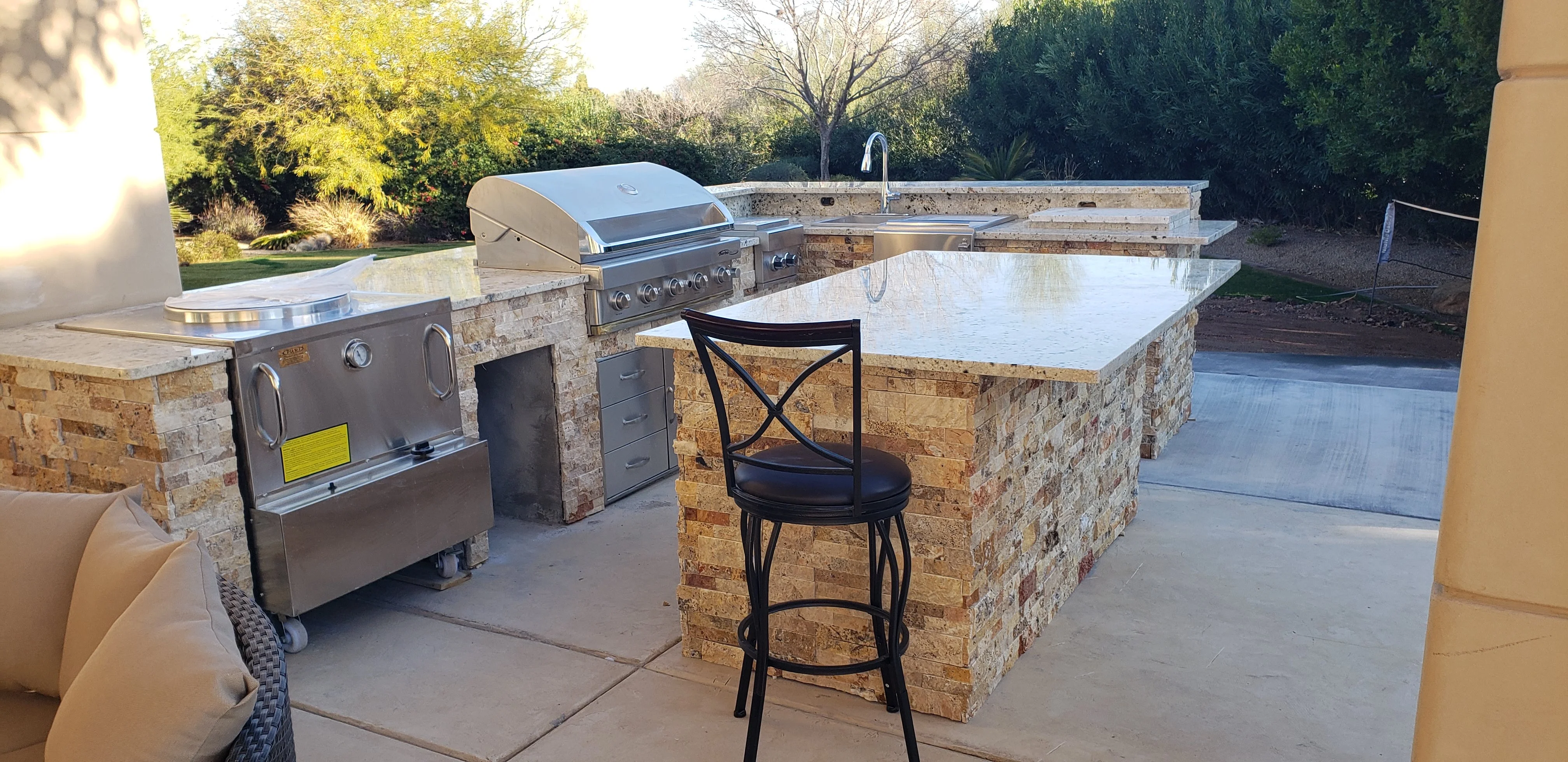 A fully equipped outdoor kitchen with stainless steel grill, sink, and storage cabinets built into a stone countertop island on a concrete patio.