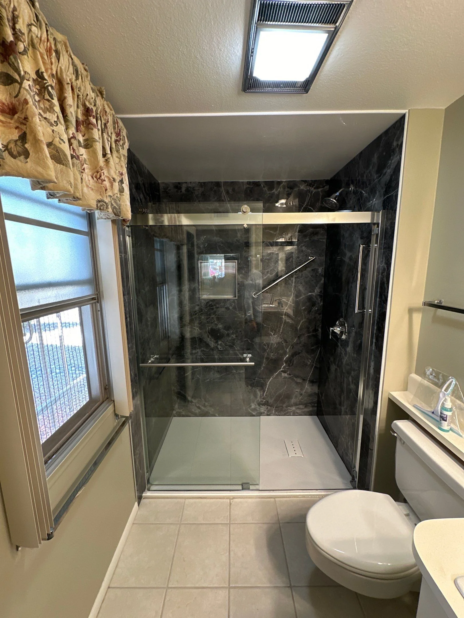 Modern walk-in shower with dark marble-patterned wall panels and sliding glass doors next to a window
