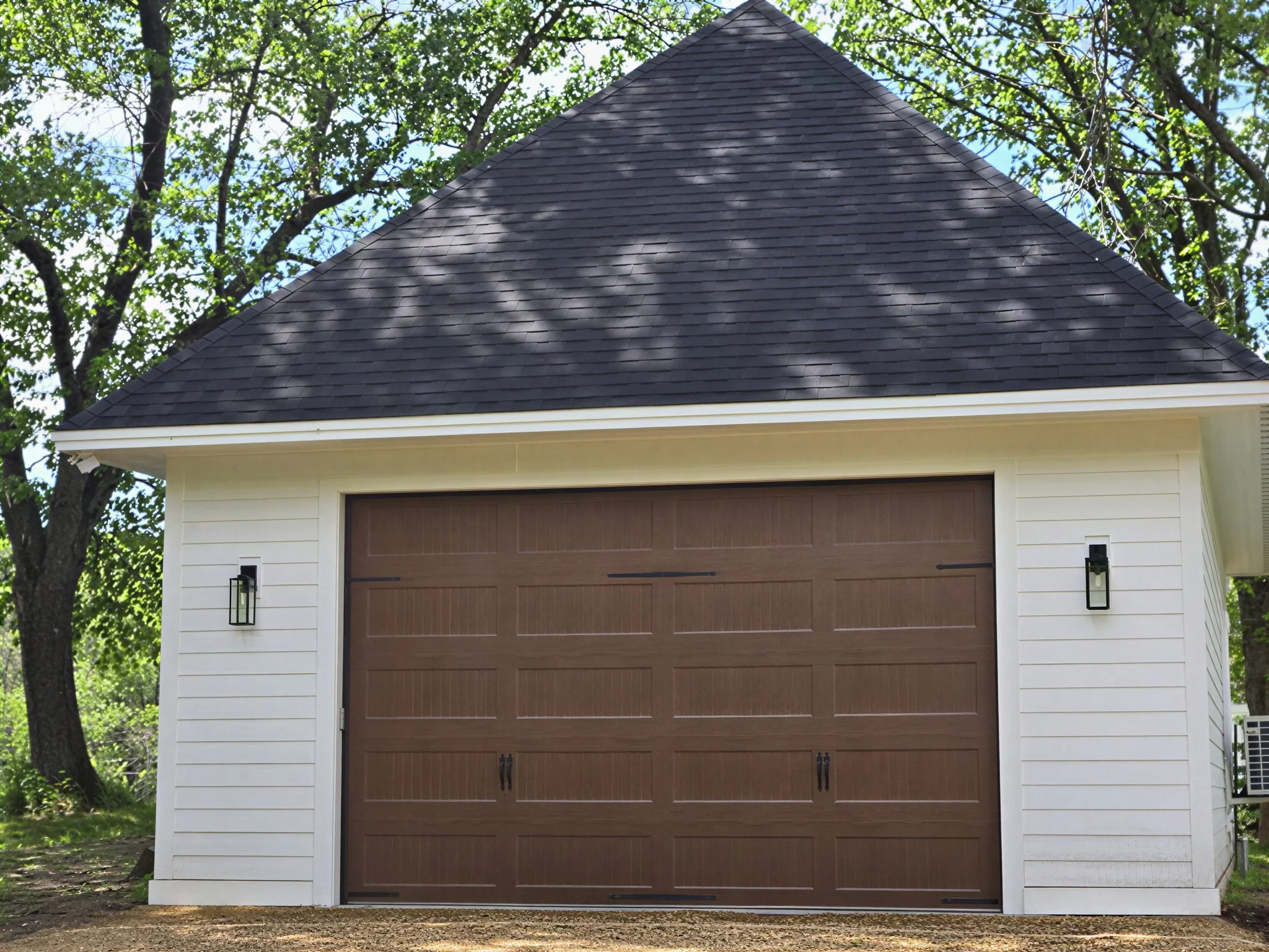 Modern white detached garage with dark hip roof, brown carriage-style door, and black sconces.