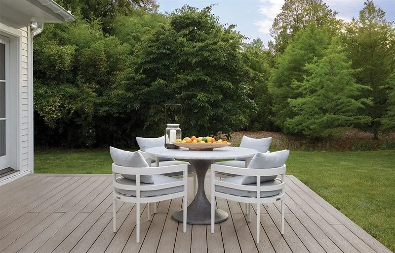 Modern white outdoor dining set on a gray composite deck with lush green tree backdrop