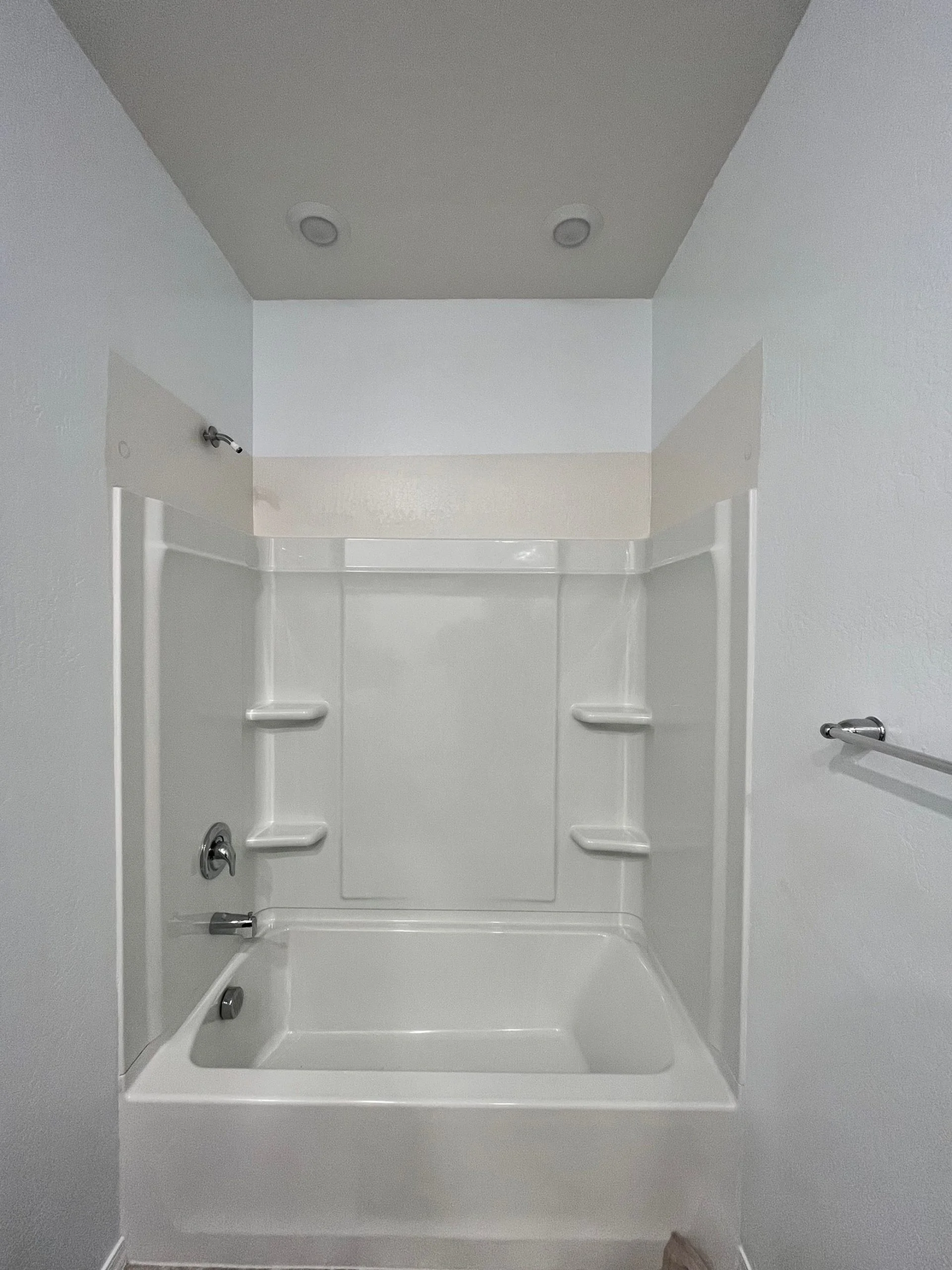 White acrylic bathtub and shower surround with built-in shelving and chrome fixtures in a bathroom.