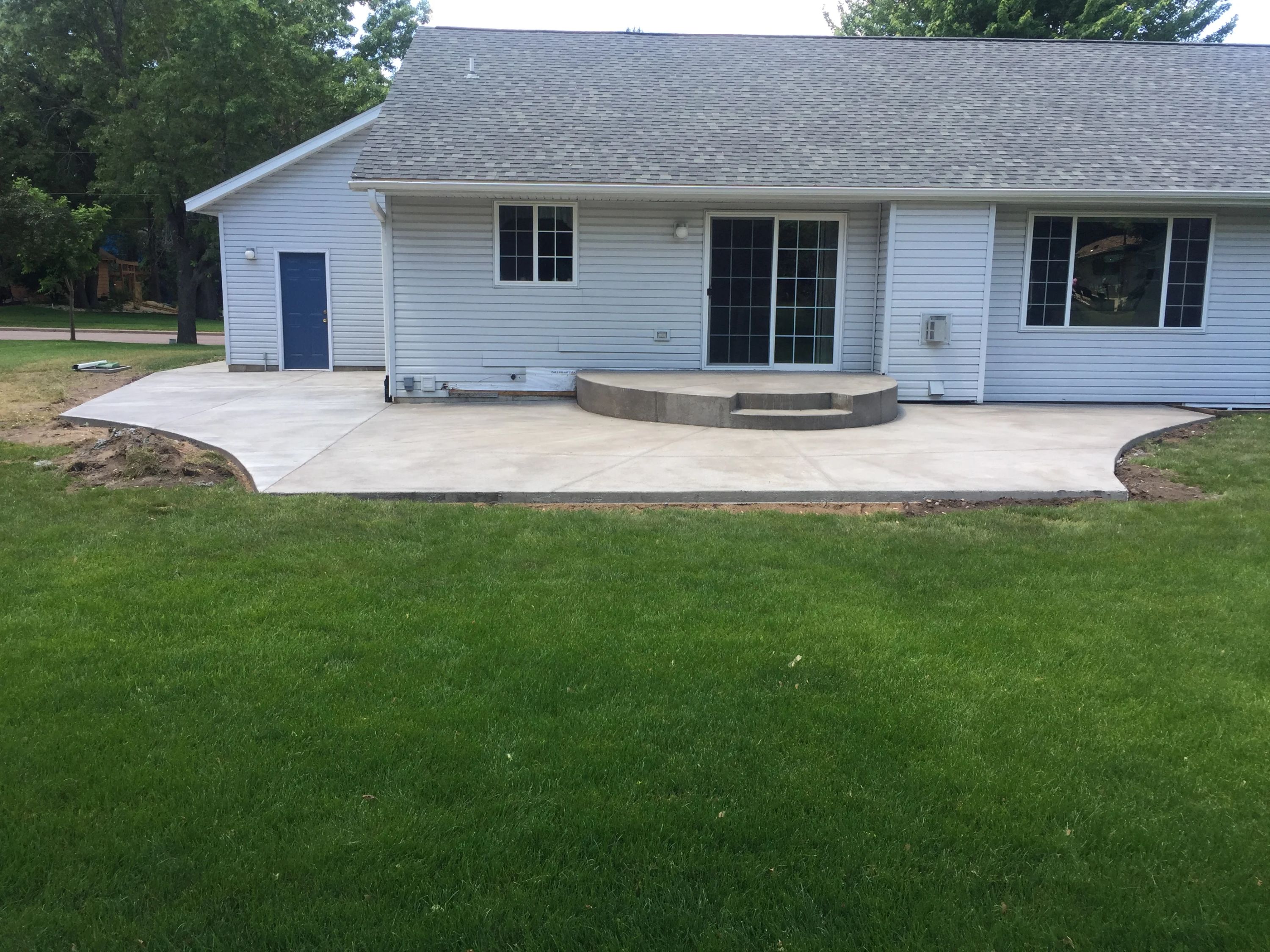 A concrete patio with curved seating wall installed behind a white single-story residential home with multiple windows and glass doors.