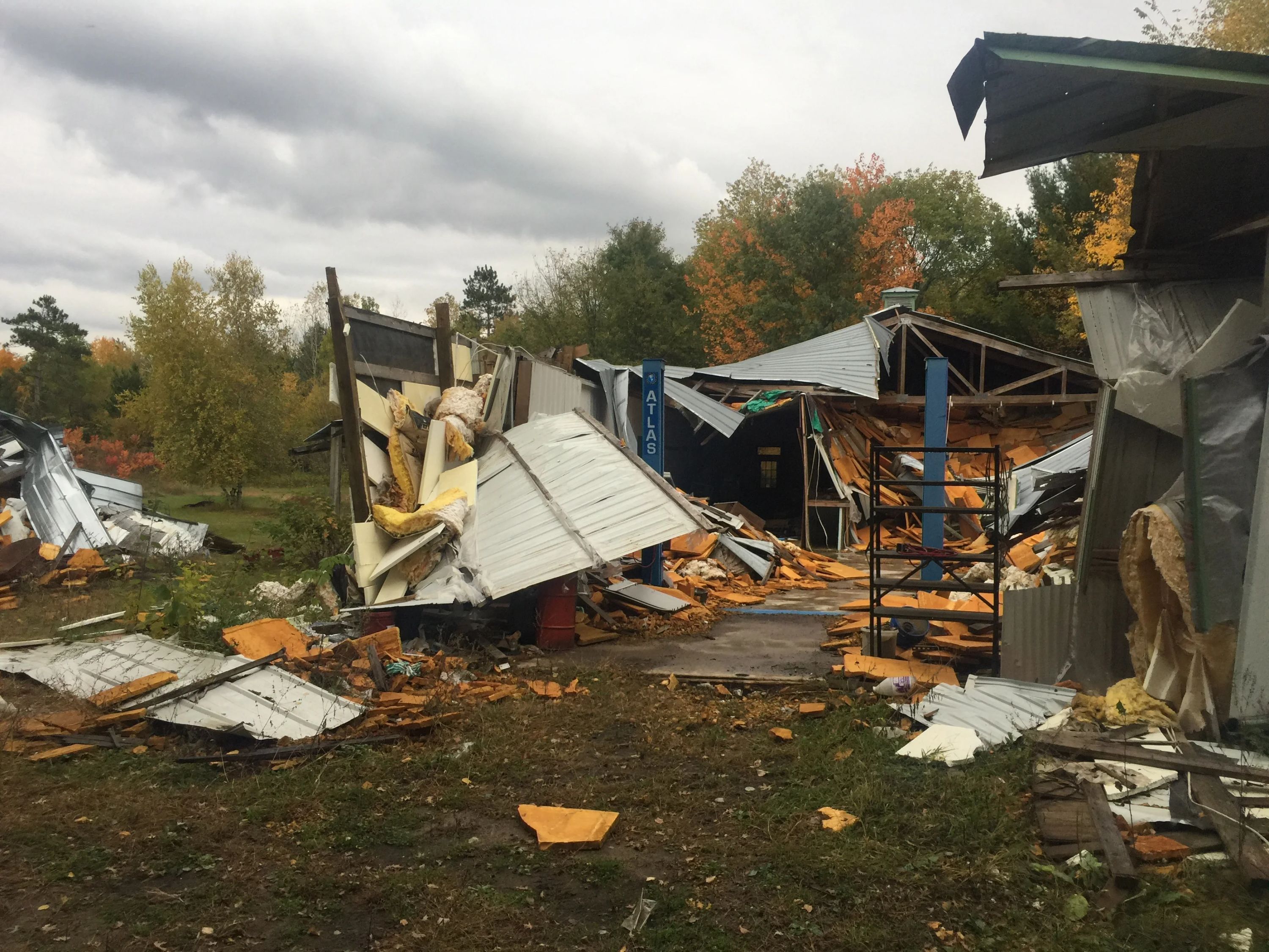 Salvage yard with discarded metal roofing, structural materials, and industrial debris scattered across a dirt lot surrounded by autumn trees.