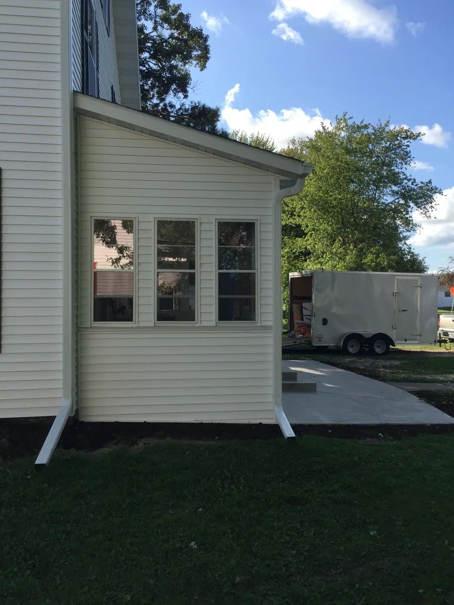 White vinyl-sided home addition with multi-pane windows and sliding glass door opening onto a concrete patio in a residential backyard setting.